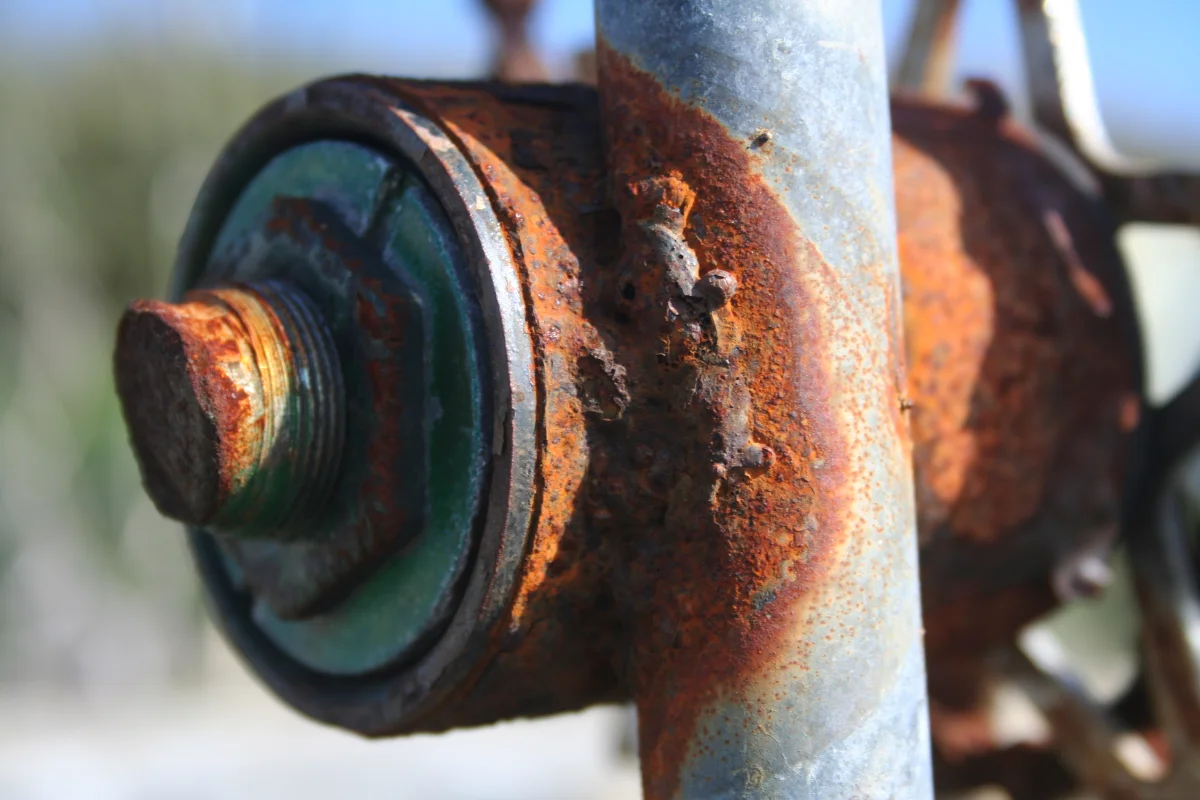 Close-up photograph of a heavily rusted iron bolt and metal fitting showing orange-brown iron oxide (rust) and green patina from galvanic corrosion between dissimilar metals