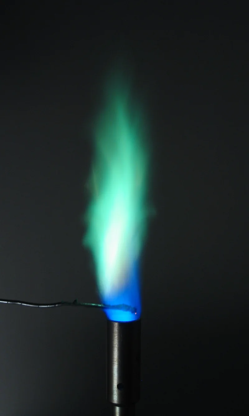 Flame test photograph showing a vivid blue-green flame produced by copper, with a Bunsen burner and nichrome wire loop visible