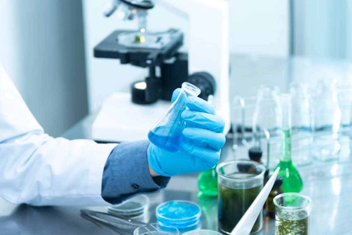Researcher in a chemistry laboratory holding an Erlenmeyer flask containing blue solution, with various reagents, beakers, and a microscope on the lab bench